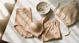 A soft flat lay of folded nude shapewear and lingerie pieces arranged on a white marble surface with a mesh laundry bag and lavender sprig, bathed in warm natural light.