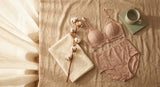 A soft flatlay of postpartum lingerie essentials including a nursing bralette and high-waisted brief arranged on warm linen in gentle morning light.