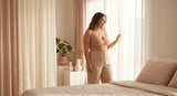 A confident woman in a well-fitted nude bralette stands in soft natural light near a window, radiating comfort and body confidence in a warm, minimal bedroom setting.
