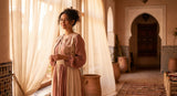 A confident Moroccan woman in an elegant, flowing outfit stands in warm golden morning light near a sunlit window with soft Moroccan architectural details in the background.